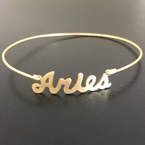 May include: Gold-toned bangle bracelet with the word "Aries" written in a cursive font.