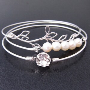 May include: A set of three silver-toned bangles. One bangle features a clear, faceted gemstone. Another has a decorative branch with leaves and five white pearls. The bangles are on a dark surface.
