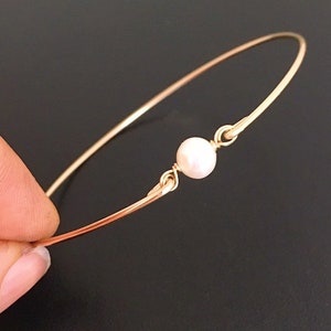 May include: A delicate gold bangle bracelet with a single white pearl accent.