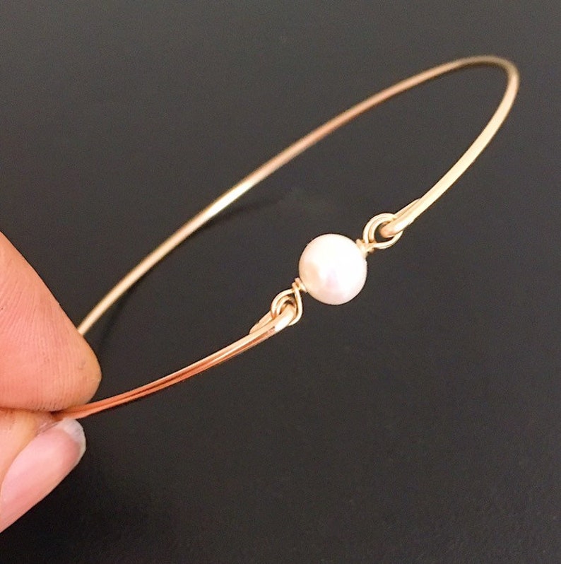 May include: A delicate gold bangle bracelet with a single white pearl accent.