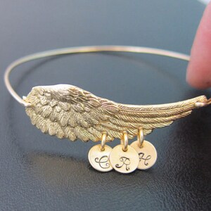 May include: Gold bangle bracelet with a detailed angel wing charm and three personalized charms with the letters C, R, and H.