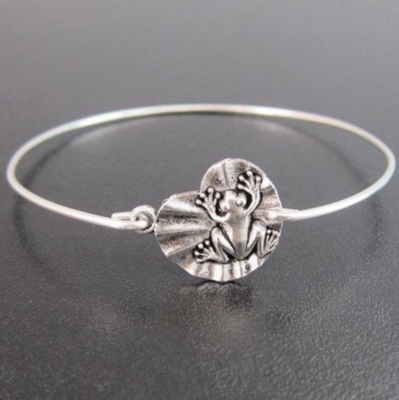 May include: A silver bangle bracelet with a frog charm. The frog is sitting on a lily pad.