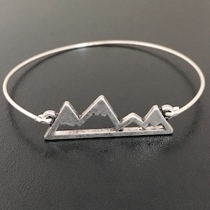 May include: Silver-toned bangle bracelet featuring a mountain range design. The bracelet has a thin, circular band with a detailed mountain silhouette in the center. The mountains are a matte silver color, and the bracelet is set against a dark background.