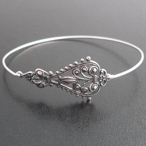 May include: A silver-toned bangle bracelet with an ornate filigree design. The central decorative element features intricate scrollwork and circular accents. The bracelet has a delicate, open design, suitable for everyday wear or special occasions.