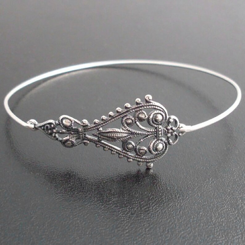 May include: A silver-toned bangle bracelet with an ornate filigree design. The central decorative element features intricate scrollwork and circular accents. The bracelet has a delicate, open design, suitable for everyday wear or special occasions.
