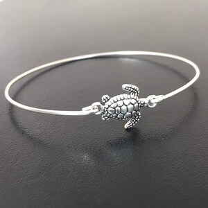 May include: A silver bangle bracelet with a turtle charm. The turtle is detailed with a textured shell and is facing to the right.