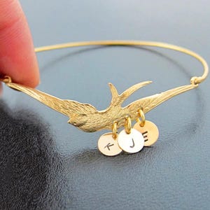 Peut inclure: Bracelet jonc dor&eacute; avec un motif d'oiseau aux ailes d&eacute;ploy&eacute;es. Trois petits charms ronds sont suspendus en dessous, chacun grav&eacute; d'une lettre. Le bracelet est tenu par une main sur un fond sombre.