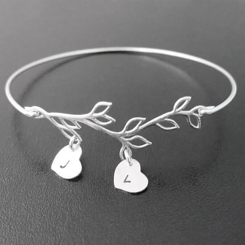 May include: Silver bangle bracelet with a delicate branch design and two dangling heart charms. Each heart charm has a letter engraved on it.