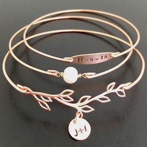 K&ouml;nnte beinhalten: Ein Set aus drei ros&eacute;goldenen Armreifen mit einem zarten Zweig-Anh&auml;nger und einem kleinen runden Anh&auml;nger mit den Initialen "J+I". Der mittlere Armreif hat eine kleine Perle und ein rechteckiges Schild mit der eingravierten Jahreszahl "11-9-19".
