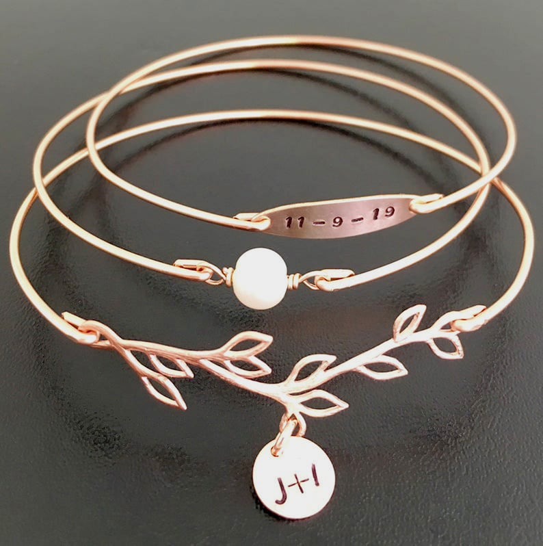 K&ouml;nnte beinhalten: Ein Set aus drei ros&eacute;goldenen Armreifen mit einem zarten Zweig-Anh&auml;nger und einem kleinen runden Anh&auml;nger mit den Initialen "J+I". Der mittlere Armreif hat eine kleine Perle und ein rechteckiges Schild mit der eingravierten Jahreszahl "11-9-19".