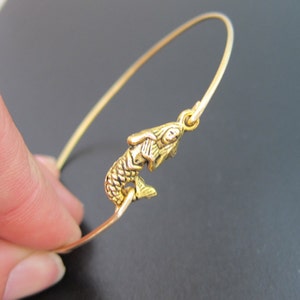 Puede incluir: Un brazalete de alambre dorado con un peque&ntilde;o dije de pez detallado. El dije est&aacute; orientado hacia arriba y la boca del pez est&aacute; abierta.