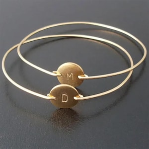 May include: Two gold bangle bracelets with round charms. The charms have the letters "M" and "D" engraved on them.
