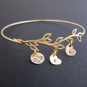 May include: Gold bangle bracelet with a delicate branch design and three charms. The charms are a heart with the letter "E", a circle with "D+B", and a heart with the letter "S".