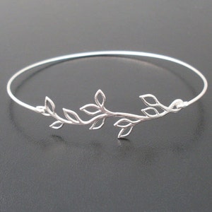 May include: A silver-colored bangle bracelet featuring a delicate leaf design. The leaves are intricately detailed and arranged along a slender, circular band. The bracelet is shown against a dark background.