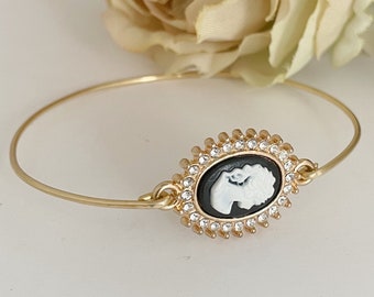 May include: A gold bangle bracelet with a black and white cameo featuring a woman's profile. The cameo is surrounded by a circle of clear rhinestones.