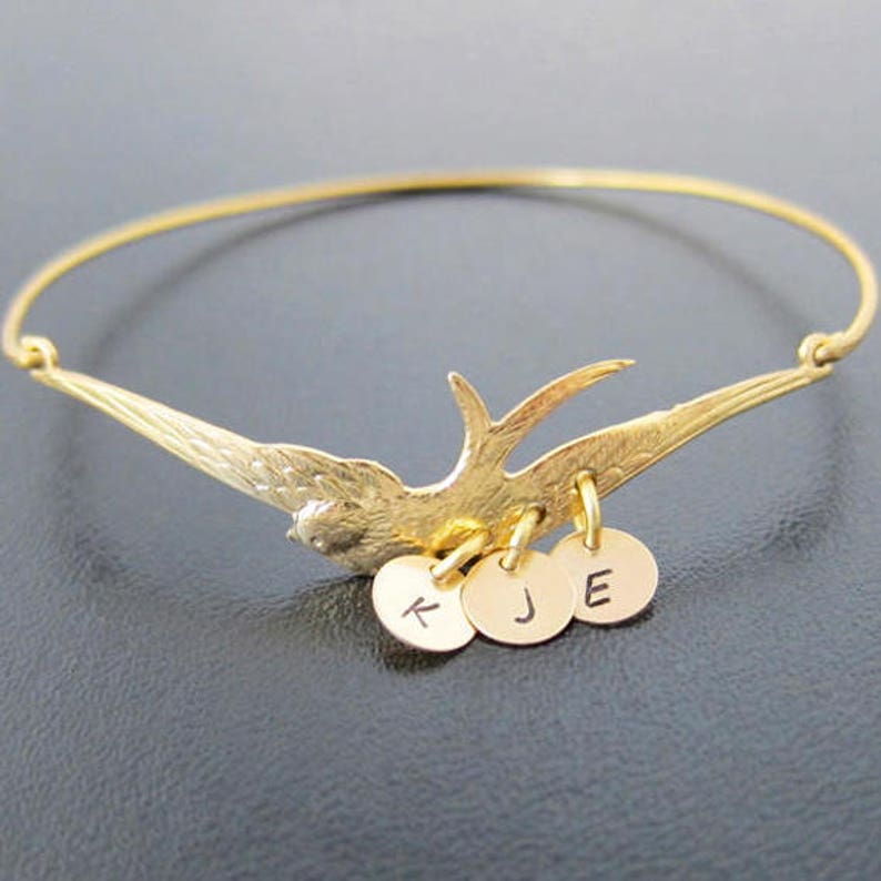 Peut inclure: Bracelet jonc dor&eacute; avec un motif d'oiseau aux ailes d&eacute;ploy&eacute;es. Trois petits charms ronds, chacun avec une lettre, sont suspendus en dessous. Le bracelet est pos&eacute; sur un fond sombre.