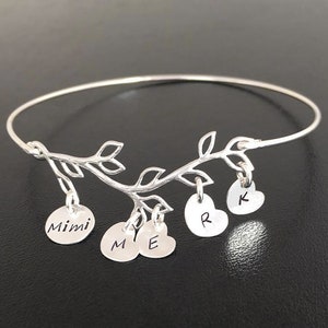 May include: Silver bangle bracelet with a delicate branch design and five charms. The charms are round and heart-shaped with the names "Mimi", "M", "E", "R", and "K" engraved on them.
