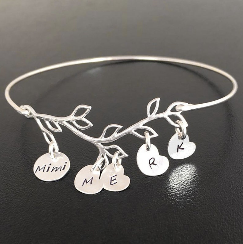May include: Silver bangle bracelet with a delicate branch design and five charms. The charms are round and heart-shaped with the names "Mimi", "M", "E", "R", and "K" engraved on them.