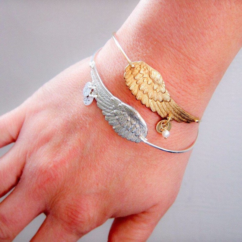 May include: A silver and gold bangle bracelet with two angel wing charms. The silver wing charm has a small charm with a pearl on it. The gold wing charm has a small charm with a letter on it.