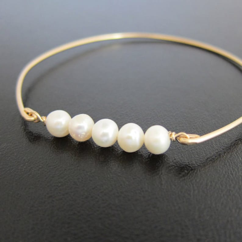 K&ouml;nnte beinhalten: Ein goldfarbenes Armreif-Armband mit f&uuml;nf wei&szlig;en Perlen. Die Perlen sind gleichm&auml;&szlig;ig &uuml;ber die Vorderseite des Armbands verteilt. Das Armband hat ein schlichtes, elegantes Design mit glatter, polierter Oberfl&auml;che.