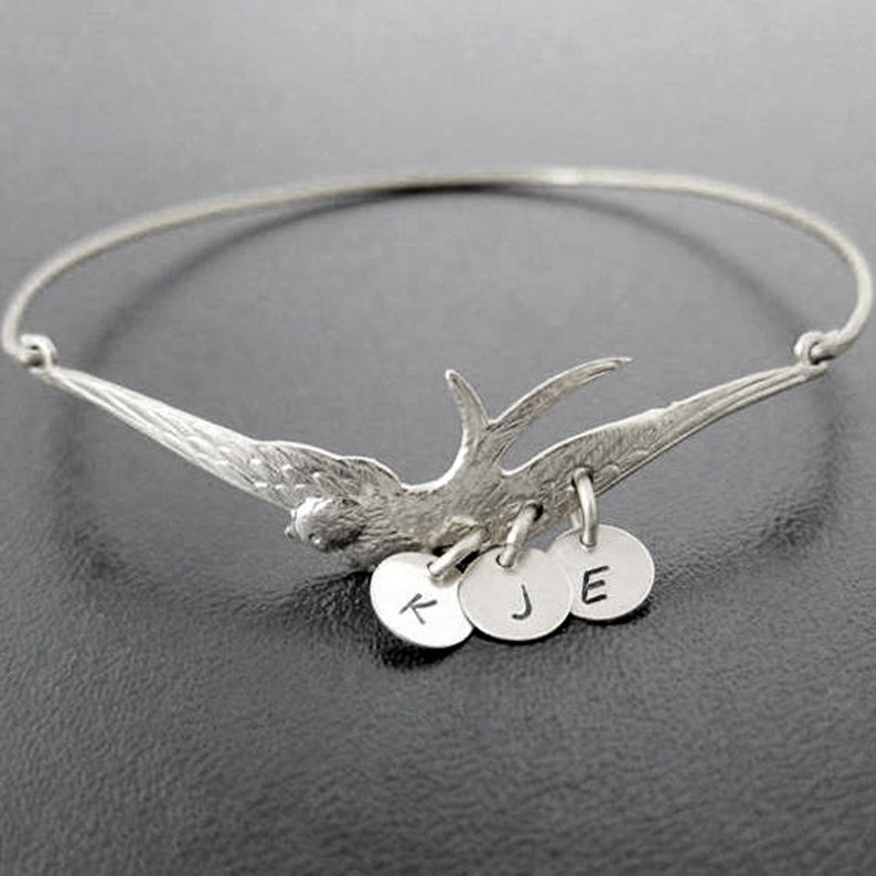 Peut inclure: Un bracelet jonc argent&eacute; orn&eacute; d'un motif d'oiseau d&eacute;taill&eacute;. L'oiseau a des ailes d&eacute;ploy&eacute;es et est accompagn&eacute; de trois petits charms ronds, chacun grav&eacute; d'une lettre. Le bracelet est pr&eacute;sent&eacute; sur un fond sombre.