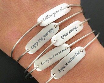May include: Five silver bangle bracelets with engraved inspirational messages: "Follow your bliss", "Enjoy the journey", "Live your dream", "Grow strong", and "Expect miracles".