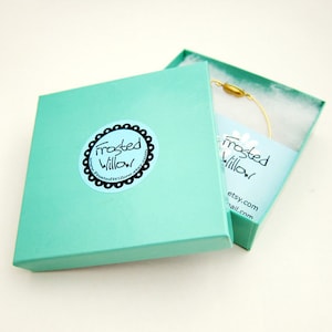 Puede incluir: Una caja de regalo turquesa con una etiqueta blanca que dice "Frosted Willow" y una direcci&oacute;n de sitio web. La caja est&aacute; abierta y un brazalete de alambre dorado est&aacute; dentro.