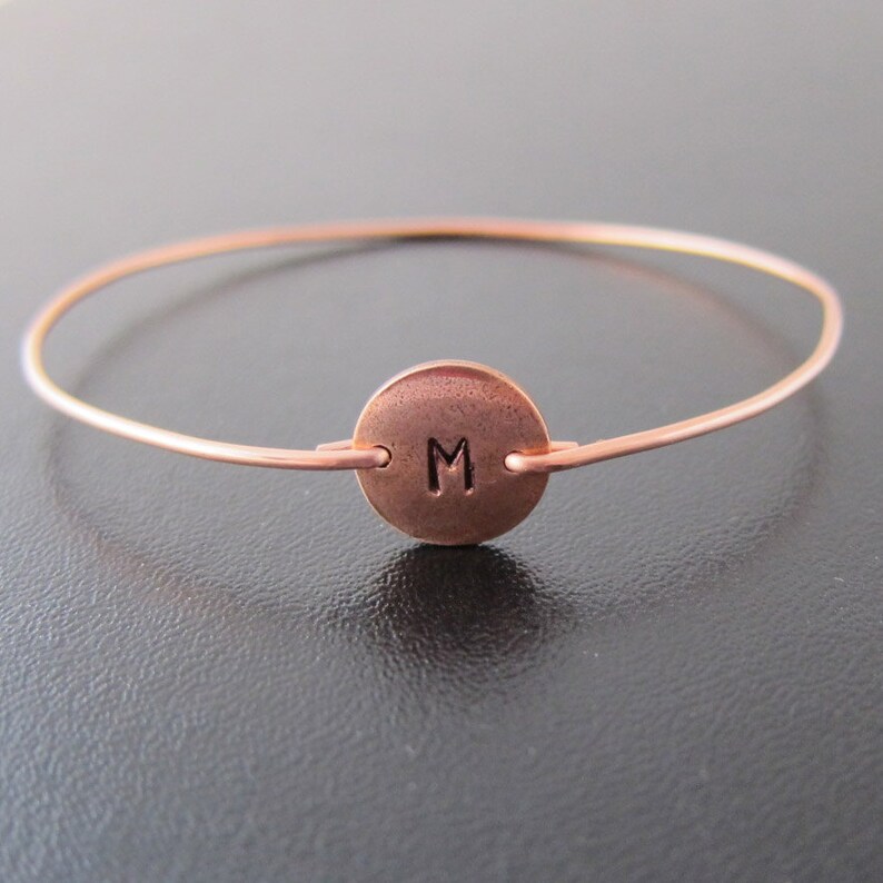 K&ouml;nnte beinhalten: Ein kupferfarbenes Armreif-Armband mit einem kreisf&ouml;rmigen Anh&auml;nger. Der Anh&auml;nger zeigt den Buchstaben "M" in einer einfachen Schriftart. Das Armband besteht aus d&uuml;nnem Draht und hat ein minimalistisches Design, geeignet f&uuml;r den Alltag. Der Durchmesser betr&auml;gt ca. 6 cm.