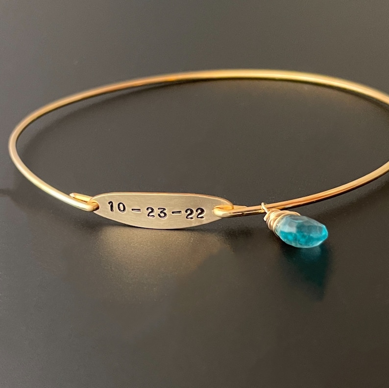 Pu&ograve; includere: Braccialetto rigido in oro con un piccolo ciondolo in pietra turchese e un'etichetta incisa con la data "10-23-22".