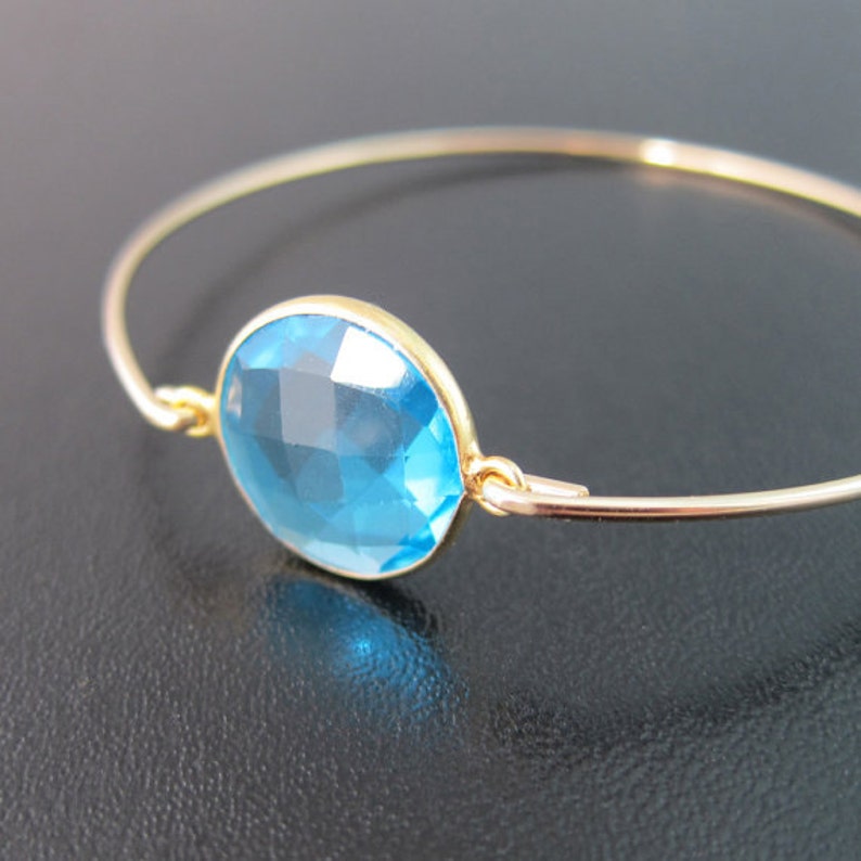 May include: A gold bangle bracelet featuring a large, light blue gemstone. The gemstone is faceted and has a round shape.