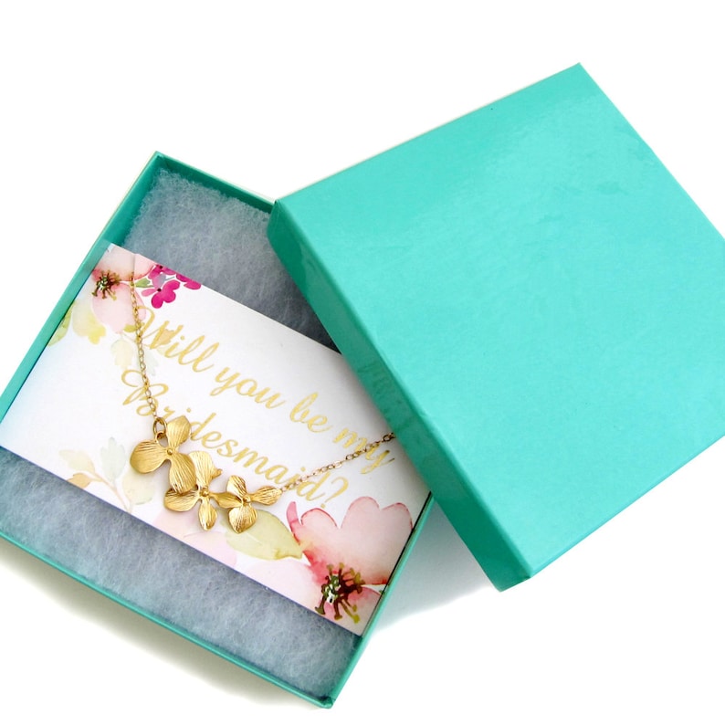 May include: A turquoise gift box with a white card inside that says "Will you be my bridesmaid?"  A gold necklace with a flower pendant is in the box.