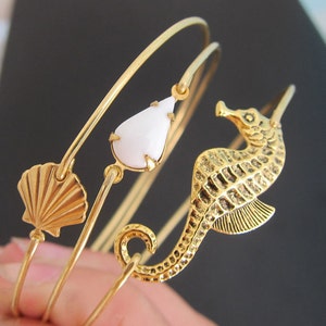 Pode incluir: Conjunto de pulseiras de bracelete douradas com um amuleto de l&aacute;grima branca, um amuleto de concha dourada e um amuleto de cavalo-marinho dourado.