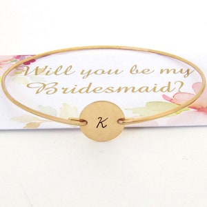 Puede incluir: Pulsera de oro con un dije redondo grabado con la letra K. La pulsera est&aacute; sobre una tarjeta que dice "&iquest;Quieres ser mi dama de honor?"