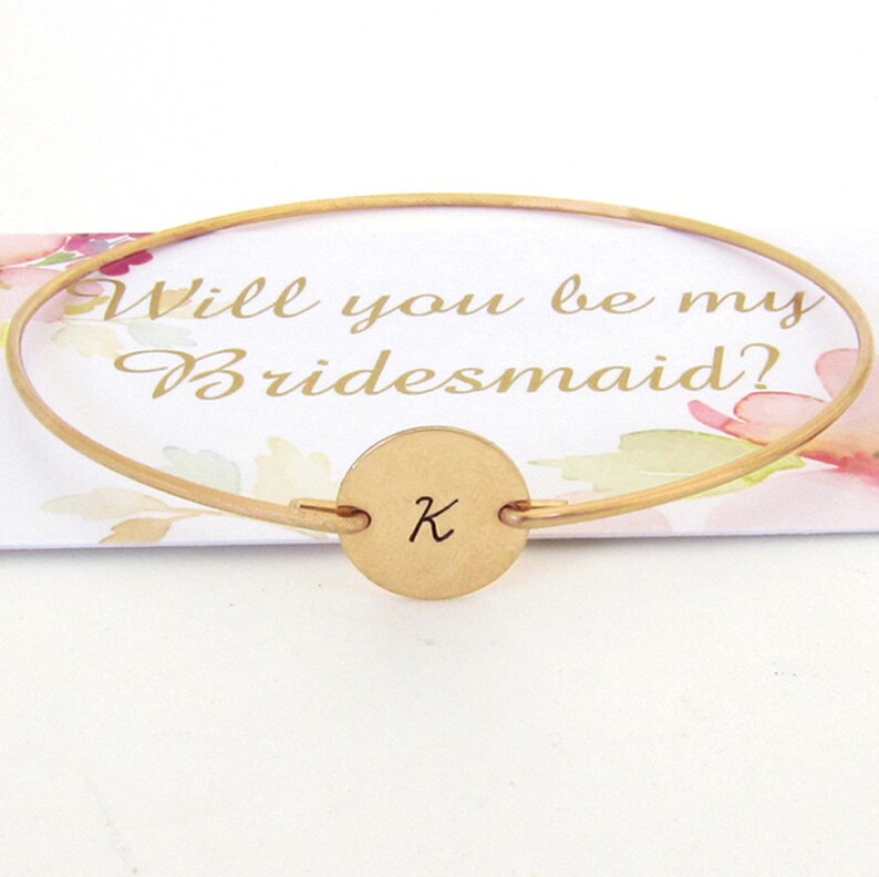 Puede incluir: Pulsera de oro con un dije redondo grabado con la letra K. La pulsera est&aacute; sobre una tarjeta que dice "&iquest;Quieres ser mi dama de honor?"