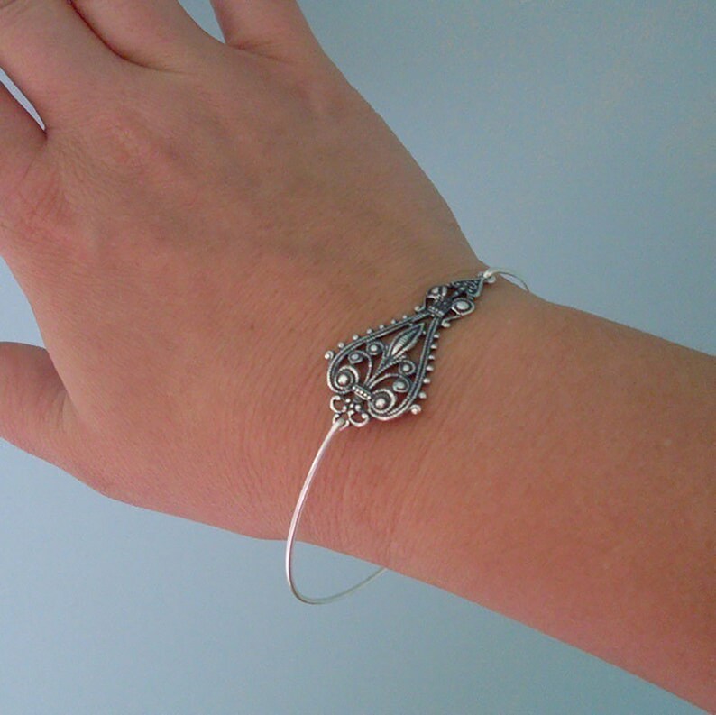 May include: A silver-toned bracelet with an ornate, teardrop-shaped centerpiece. The centerpiece features intricate filigree and scrollwork designs. The bracelet is a delicate, thin wire design, and is worn on a wrist.