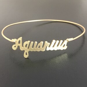 Pode incluir: Um bracelete dourado com a palavra "Aqu&aacute;rio" escrita em letra cursiva.