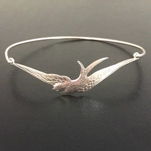 Pu&ograve; includere: Un braccialetto argentato con un design a rondine in volo. L'uccello &egrave; finemente dettagliato, con le ali spiegate. Il braccialetto &egrave; una sottile fascia di metallo curva, e la rondine &egrave; il punto focale del design.
