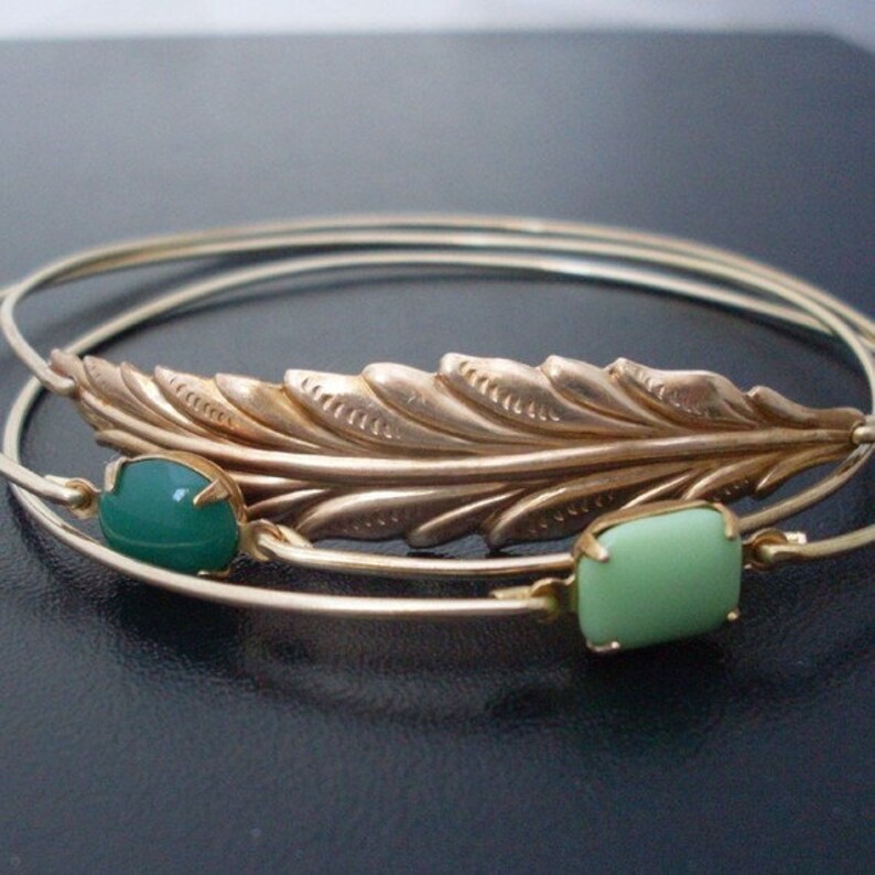 May include: Three gold bangle bracelets with a gold feather charm and two green glass stones.