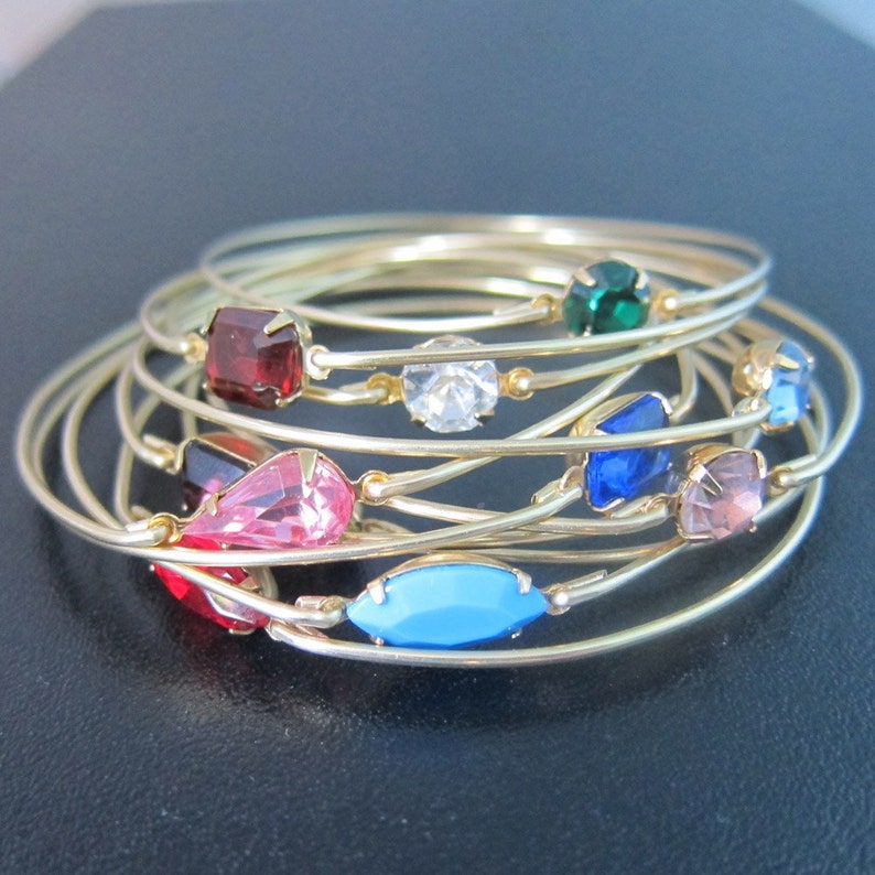 May include: A set of gold bangle bracelets with colorful rhinestones. The bracelets are stacked on top of each other and feature a variety of colors, including red, pink, blue, green, and clear.