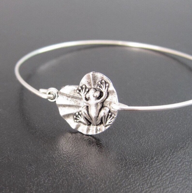 May include: A silver bangle bracelet with a silver frog charm. The frog is sitting on a lily pad.