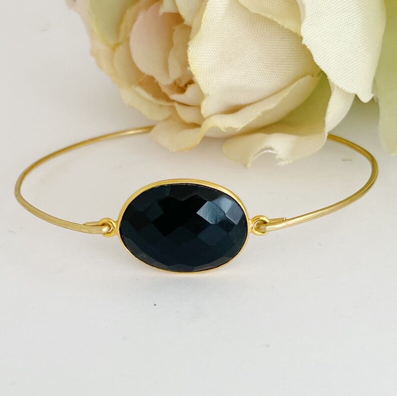 May include: A gold bangle bracelet with a black oval gemstone. The gemstone is faceted and has a high polish.