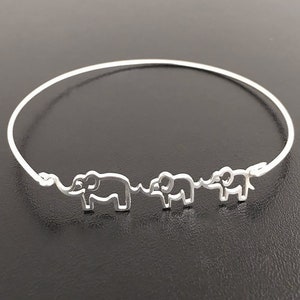 Puede incluir: Pulsera de brazalete de plata con tres siluetas de elefantes. Los elefantes est&aacute;n conectados por una l&iacute;nea fina.