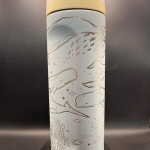 May include: A light blue insulated water bottle with a detailed etched design of whales, fish, and coral. The lid is white and the top section is yellow. The text "Lift to Unlock" is visible on the lid.