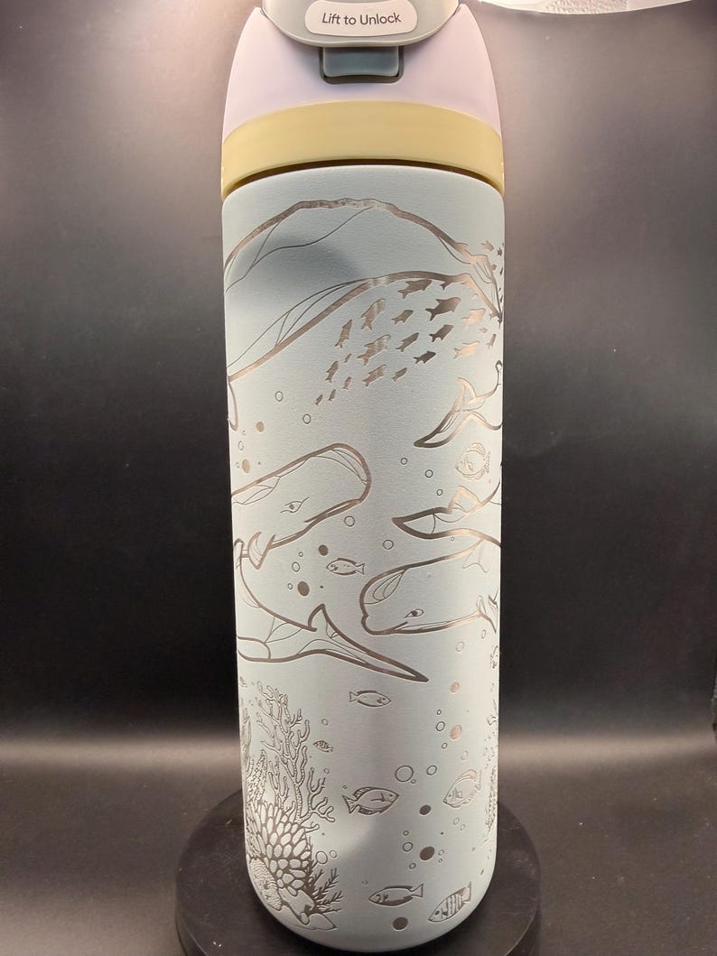 May include: A light blue insulated water bottle with a detailed etched design of whales, fish, and coral. The lid is white and the top section is yellow. The text "Lift to Unlock" is visible on the lid.