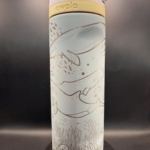 May include: A light blue Owala water bottle with a whale and coral reef design. The bottle has a light purple lid and a yellow band. The design features whales, fish, and coral in a detailed, etched style.