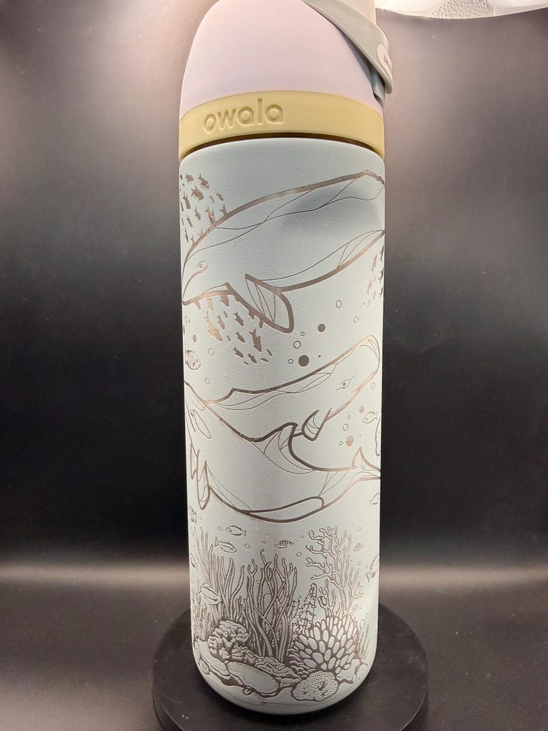May include: A light blue Owala water bottle with a whale and coral reef design. The bottle has a light purple lid and a yellow band. The design features whales, fish, and coral in a detailed, etched style.