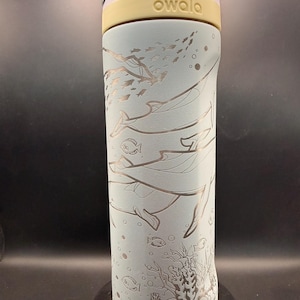 May include: A light blue Owala water bottle with a sea life design. The bottle features etched illustrations of whales, fish, and coral. The top is yellow and the lid is light purple. The bottle is cylindrical and appears to be made of metal.