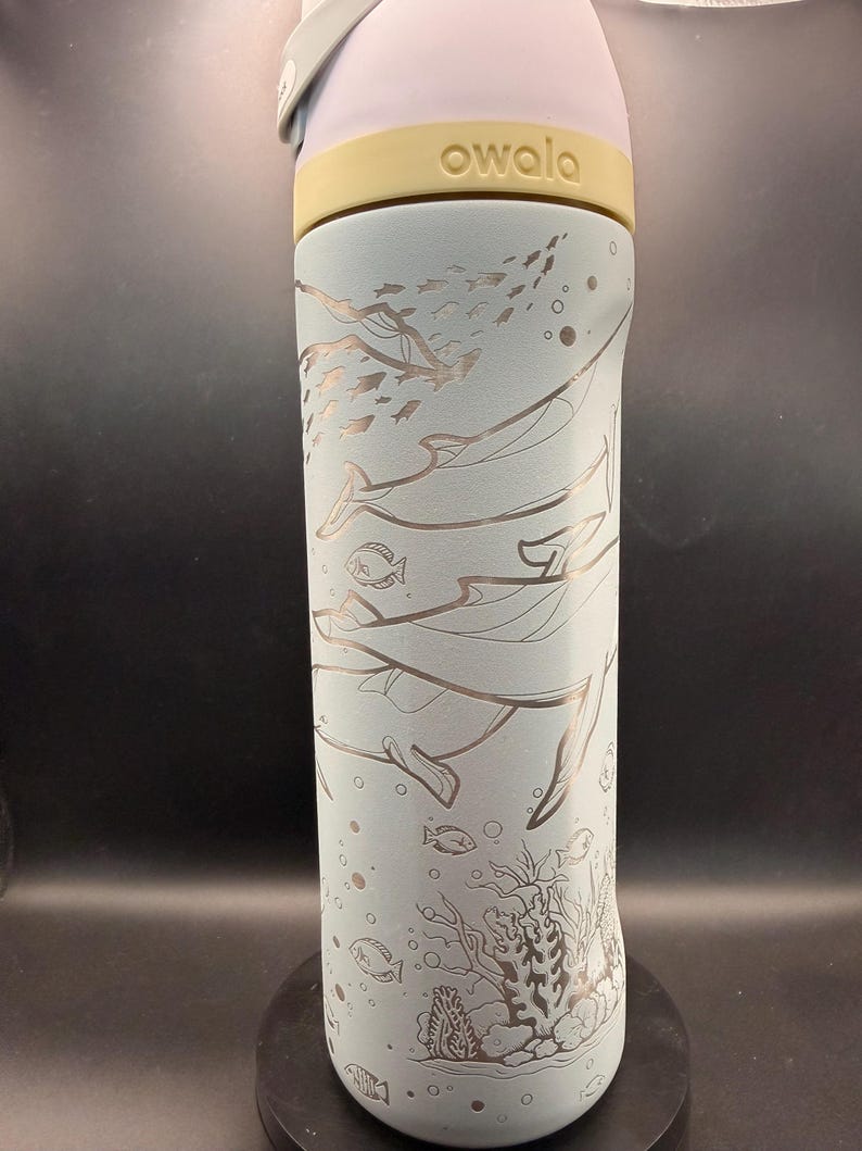 May include: A light blue Owala water bottle with a sea life design. The bottle features etched illustrations of whales, fish, and coral. The top is yellow and the lid is light purple. The bottle is cylindrical and appears to be made of metal.