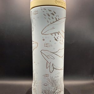 May include: A light blue Owala water bottle with a white and yellow lid. The bottle features etched illustrations of whales, fish, and coral. The word "owala" is printed on the bottle.