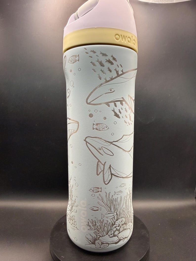 May include: A light blue Owala water bottle with a white and yellow lid. The bottle features etched illustrations of whales, fish, and coral. The word "owala" is printed on the bottle.